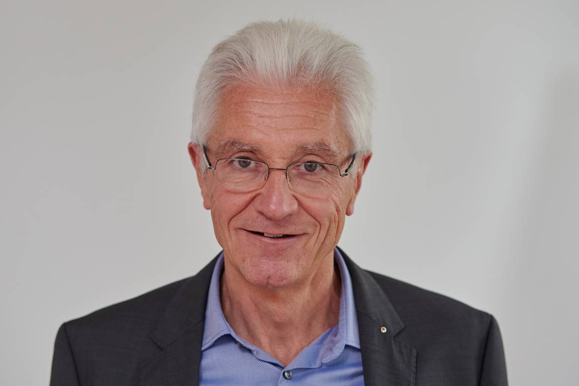 Ein älterer Mann mit weißem Haar, Brille und einer dunklen Anzugjacke über einem blauen Hemd lächelt vor einem schlichten hellen Hintergrund sanft in die Kamera.